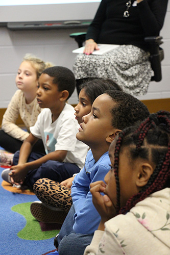 A line of five second graders sit on a colorful rug in a row watching someone who is talking to them.