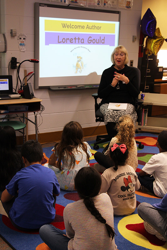 A group of second grade students sit on a colorful rug looking up at a woman with short blonde hair. The woman is reading a book to the students. Behind her is a screen that says Welcome author Loretta Gould.