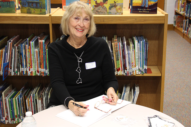 A woman with chin-length blonde hair sits at a desk and smiles. She is wearing a black long-sleeve shirt and a triple heart silver necklace. She is signing a book on the table.