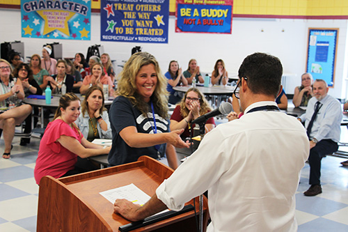 A woman with shoulder length blonde hair in a blue tshirt with white writing on it shakes hands with a man wearing a white dress shirt. He has short dark hair.