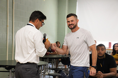 A man with short dark hair wearing a white dress shirt shakes hands with a man with dark hair, beard and mustache, wearing a white tshirt. He is smiling.