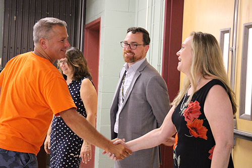 A man in an orange shirt with short light hair, shakes hands with a woman in a black dress with orange flowers on it. She has long blonde hair. There is a man next to her in a suit, with a goatee and glasses looking on.