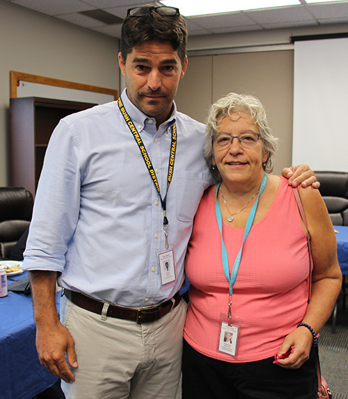 A woman with short gray hair wearing a peach colored shirt stands arm-in-arm with a man wearing a blue dress shirt and khaki pants. He has short dark hair.