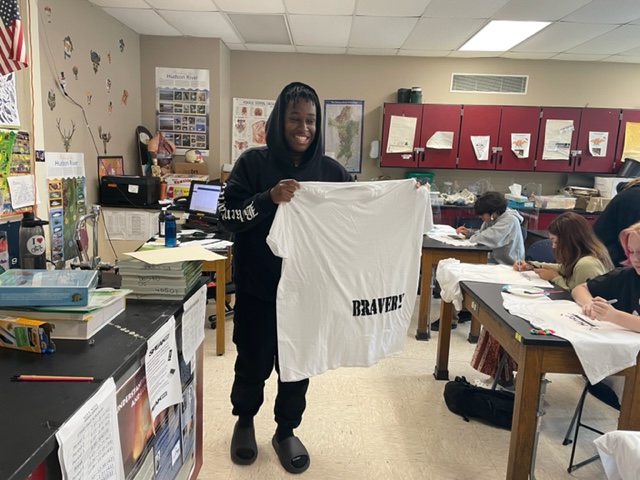 A high school age young man smiles as he holds up a white tshirt that says bravery.