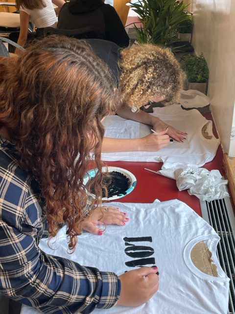 Two high school age girls  lean over a table, making tshirts. The one closest to the camera says Love.