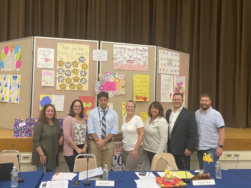 A group of adults standing behind a table. There are seven - two women, a man, two more women and two men. Behind them is a display board with many posters handmade by children. 