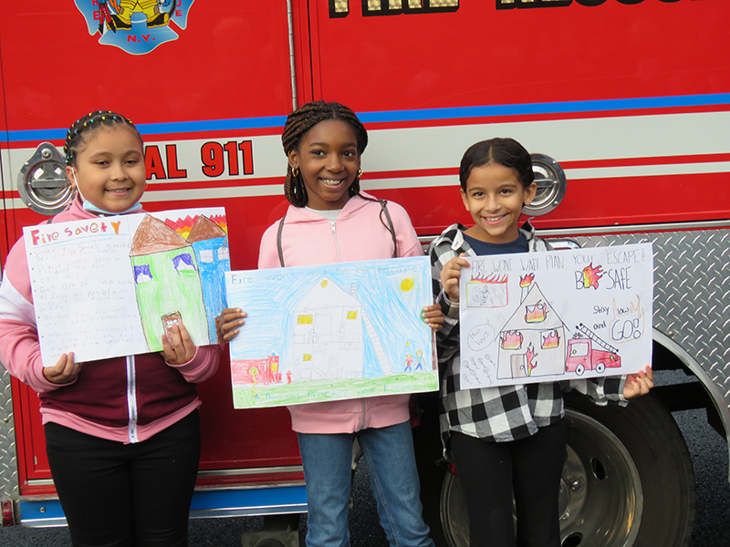 Three elementary age girls hold up posters they made for fire safety. They are standing in front of a red and white fire truck and are smiling.