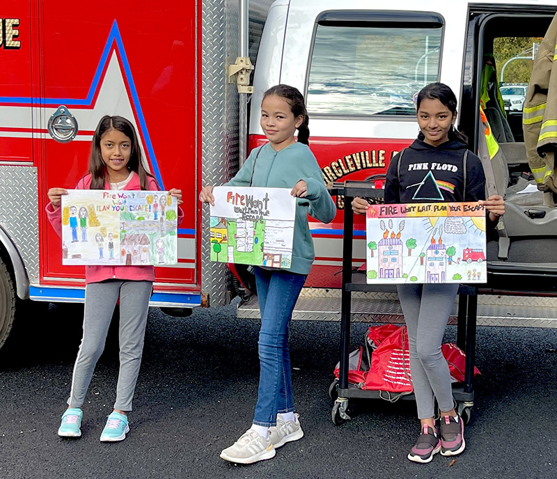 Three elementary age girls hold up posters they made for fire safety. They are standing in front of a red and white fire truck and are smiling.