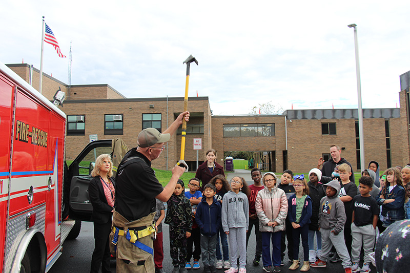 A man wearing a tan baseball cap, black tshirt, and tan firefighter pants holds up a large tool that has a metal hook on th eend of it. There is a red fire-rescue truck behind him and a group of third grade students looking up at the piece of equipment.