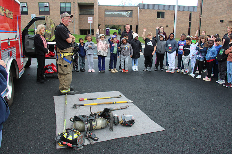 A man in a tan baseball cap, black tshirt and tan firefighter pants stands in front of a group of third grade students, some of whom have their hands raised. Next to him is equipment from a fire fighter - a helmet, an axe, an air tank and other things. Behind him is a red fire truck.