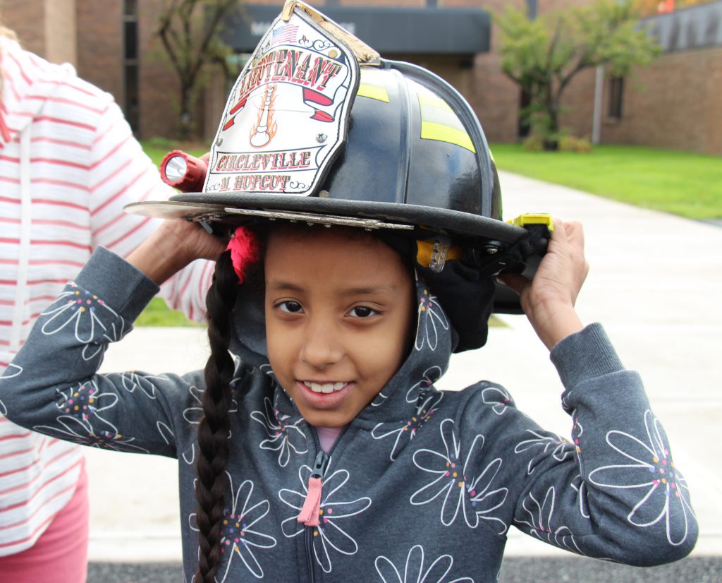 A girl with a long dark braid holds a black fire helmet on her head. She is smiling and wearing a gray sweatshirt with flowers on it.