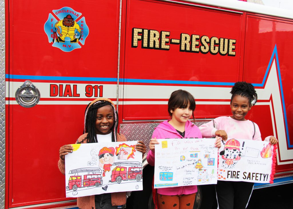 A red fire truck that says Fire-Rescue and Dial 911 is the backdrop. Three third grade girls stand in front of it, each holding a poster they made for fire safety.