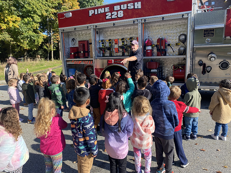 A firefighter in a navy blue shirt stands in front of a red fire truck that says Pine Bush 228 on it. He is pointing to a student with her hand up. There are lots of students in front of him.