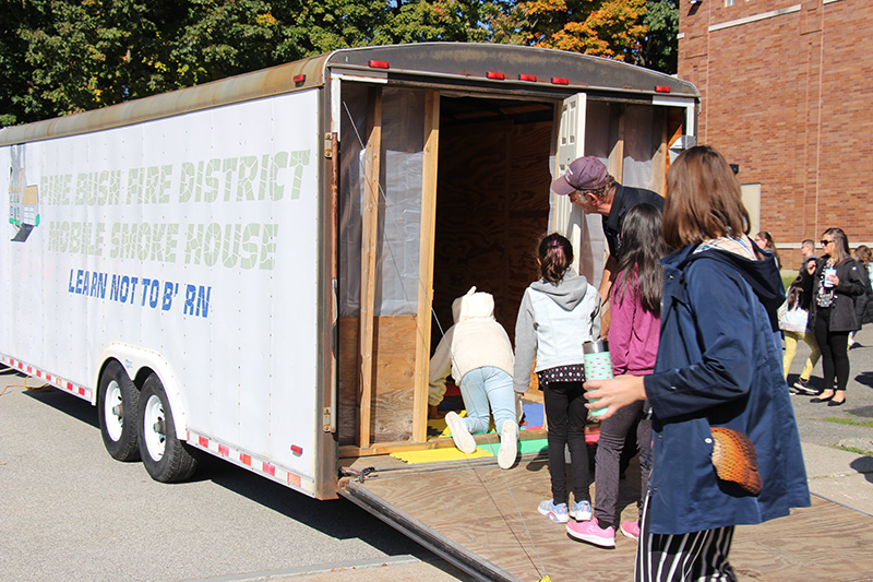 A white trailer with gold and blue letters on the side that says Pine Bush Fire district Smoke House on the side. There is a line of elementary students waiting to get in. The ones in the front are on their hands and knees to go through the smoke house.