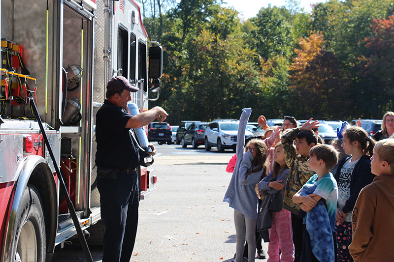 A man stands in front of a fire truck with a large group of elementary students in front of him. Some have their hands up. It's a sunny day.