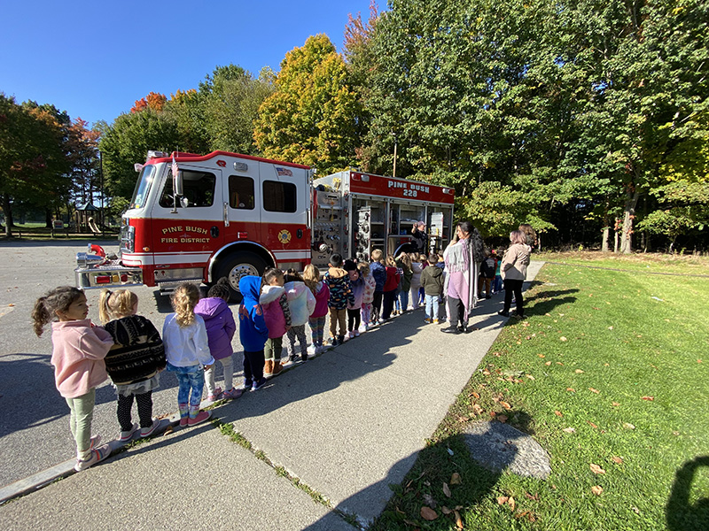 A line of pre-K students on a sidewalk stand by a red and white fire engine. It's a sunny day and the sky is blue.