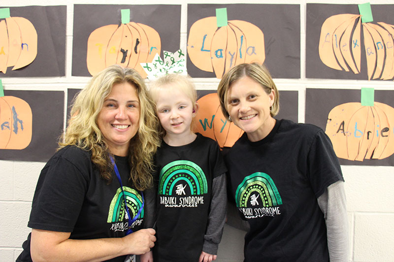 Two women smile. They are wearing black t shirts with green rainbows on them. There is a little girl in the middle wearing the same shirt and a big green and white bow in her hair. She is smiling too.