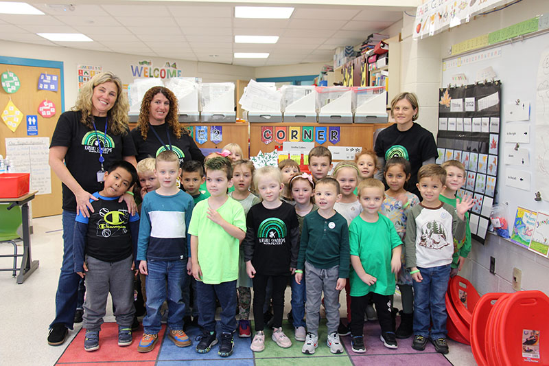 Three teachers all wearing black t shirts with green rainbows on them stand with a class of kindergarten students, about 18 of them. The kids are wearing shades of green.