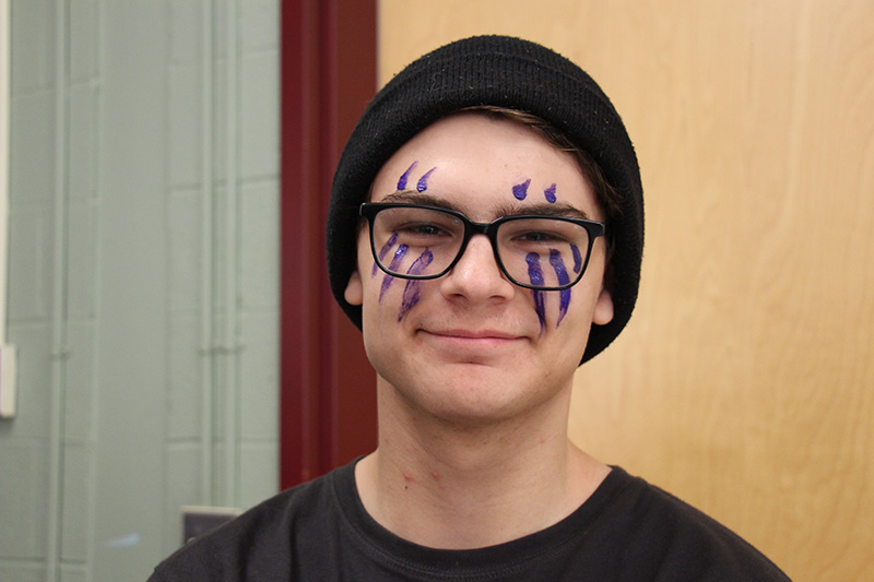 A young man wearing a black shirt and knit cap, smiles. He has glasses and purple stripes painted on his face.