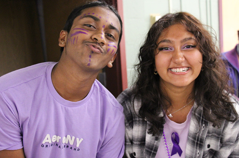 Two high school kids smile. The boy on the left is wearing a light purple shirt and has his face painted with stripes. The girl on the right has long dark hair and is wearing a purple shirt with purple ribbon and a plaid shirt over it. She is smiling broadly.