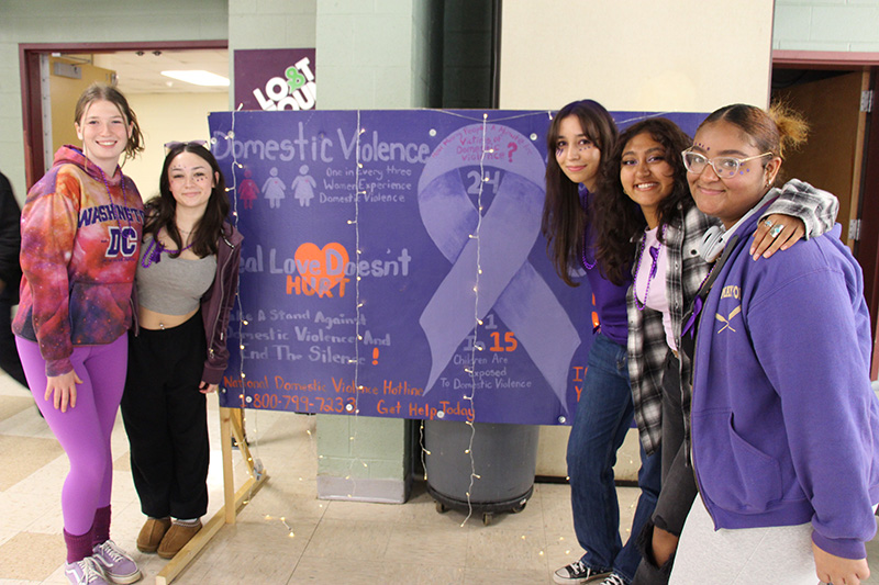 A group of five girls - two on the left and three on the right - flank a large purple sign about domestic violence.