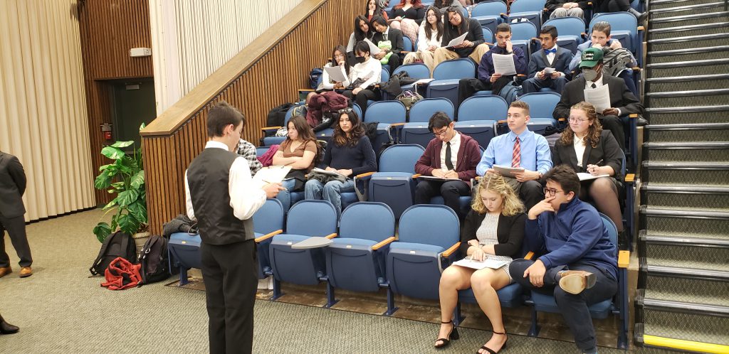 A young man in a white dress shirt and vest stands in front of a group of high school students in an auditorium. He is presenting to them.