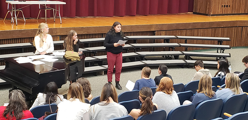 A group of high school students sit in an auditorium listening to a young woman present to them. She is holding a piece of paper in her hands and speaking to them.