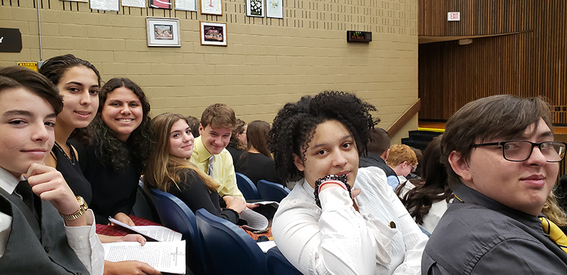 A group of seven high school students sit in an auditorium and look back at the camera.