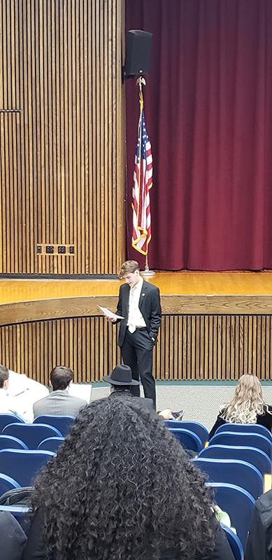 A high school student stands in front of a group of other students in an auditorium looking at a piece of paper in his hand. He is wearing a suit and tie. There is a stage behind him and an American flag on the stage. 