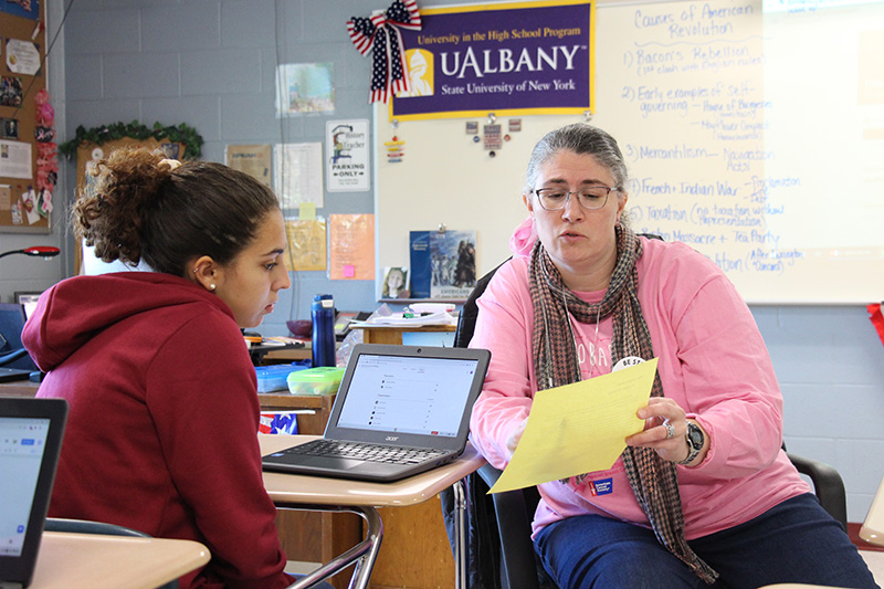 A woman wearing pink holds a piece of paper in her hand and points at words on it as she shows it to a high school student in a red hoodie. Her hair is up in a ponytail.