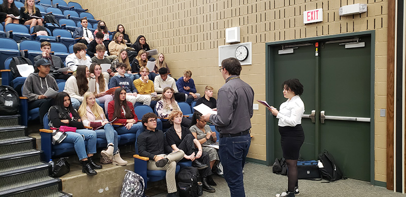 A group of high school students sit in an auditorium and listen to a high school young man talk. 