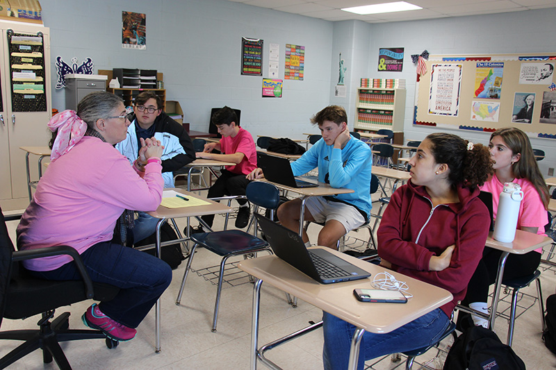 A woman wearing a pink shirt sits at a desk facing studnets in the classroom There are just five students sitting at desks gathered around her at th efront of the room.