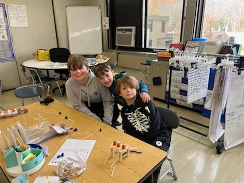 Three elementary age boys sit together with their science projects on their table with arms around each other.