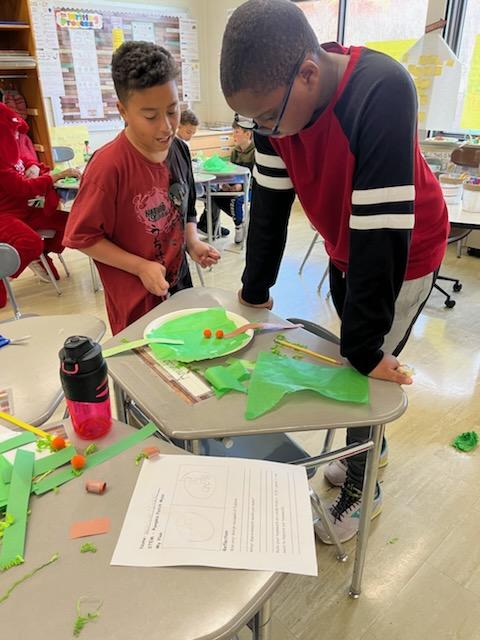 Two elementary age boys lean over a table looking at a science experiment they made with paper, sticks, colored paper.