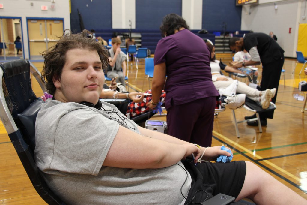 A high school boy, wearing a gray shirt, sits on a cot, holding a blue squeeze thing preparing to donate blood.