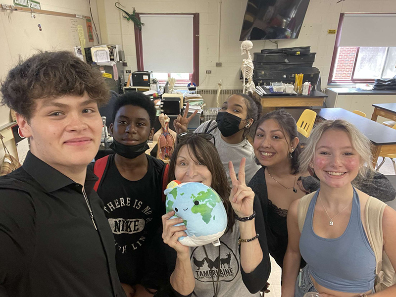 A woman is in the center, with longish dark hair, holding a pillow of the earth and a peace sign up. Around her are five high school kids all smiling.