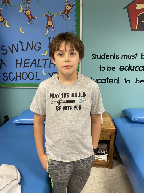 An elementary age boy, with dark hair, wears a tshirt that says May the Insulin be with you, stands