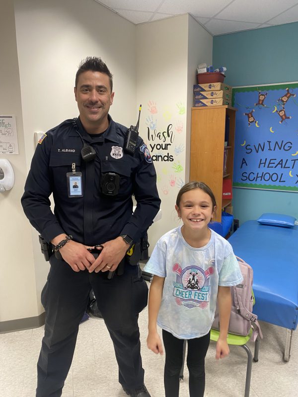 A man in a police uniform smiles and stands next to an elementary age girl wearing a light blue shirt. They are both smiling.