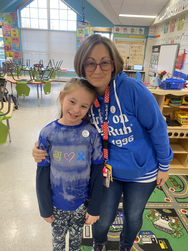 A woman with short light hair, wearing a blue sweatshirt, puts her arm around a little girl with long blonde hair pulled back, who is also wearing a blue shirt.