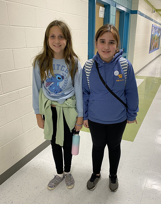 Two older elementary girls, one with long brown hair and the other with long brown hair pulled into a ponytail, stand together wearing blue.