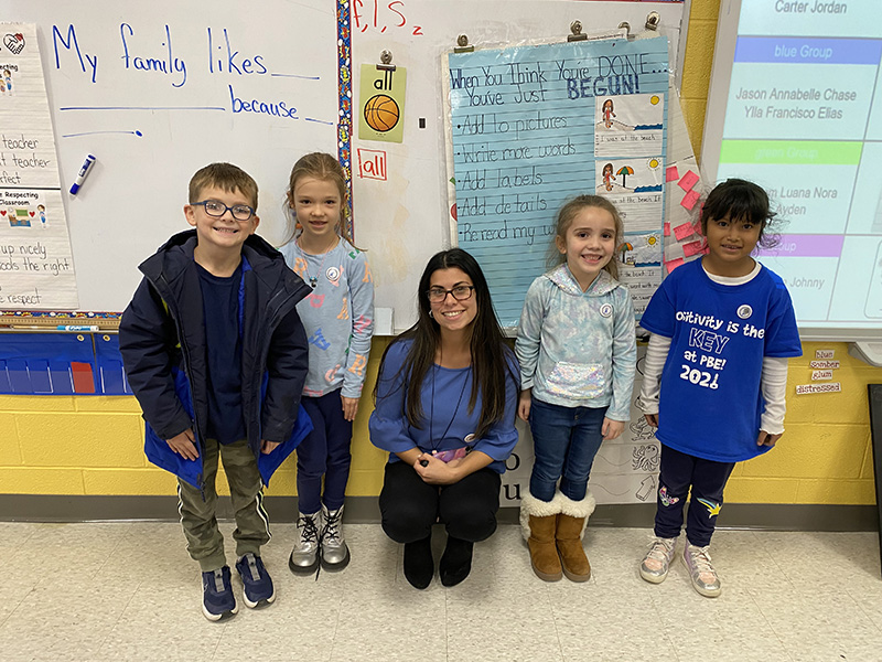 A woman with long dark hair squats in the center of four younger elementary students. All are wearing blue.