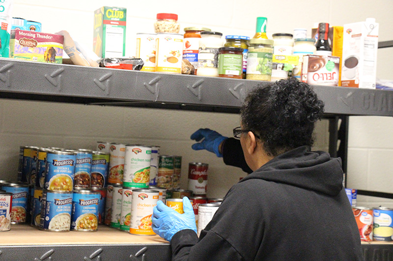 A woman with dark hair pulled up reaches to the back of a shelf filled with cans.