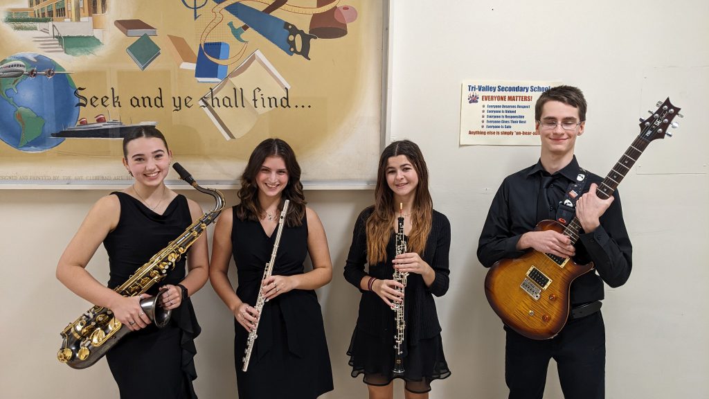 Four high school students stand smiling, all dressed in black. They are holding their musical instruments, three girls from left and a boy holding a guitar on the right.