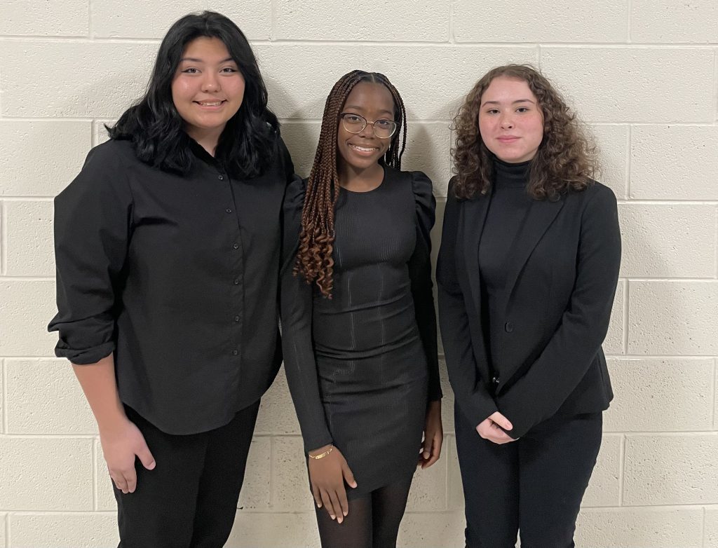 Three high school young women stand together smiling. They are all wearing black. 