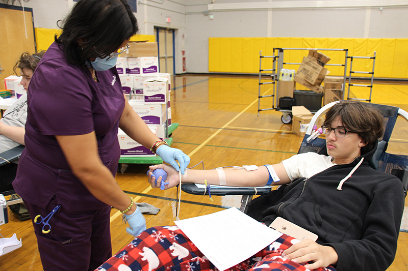 A medical technician, dressed in purple scrubs, sets up a student for giving blood. He is watching as she adjusts the line coming from his arm.