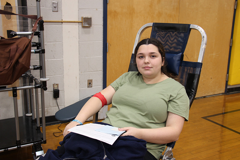 A high school girl, with dark hair, wearing a green shirt sits up on a cot. She has paperwork on her lap and a red bandage around her arm where she gave blood.