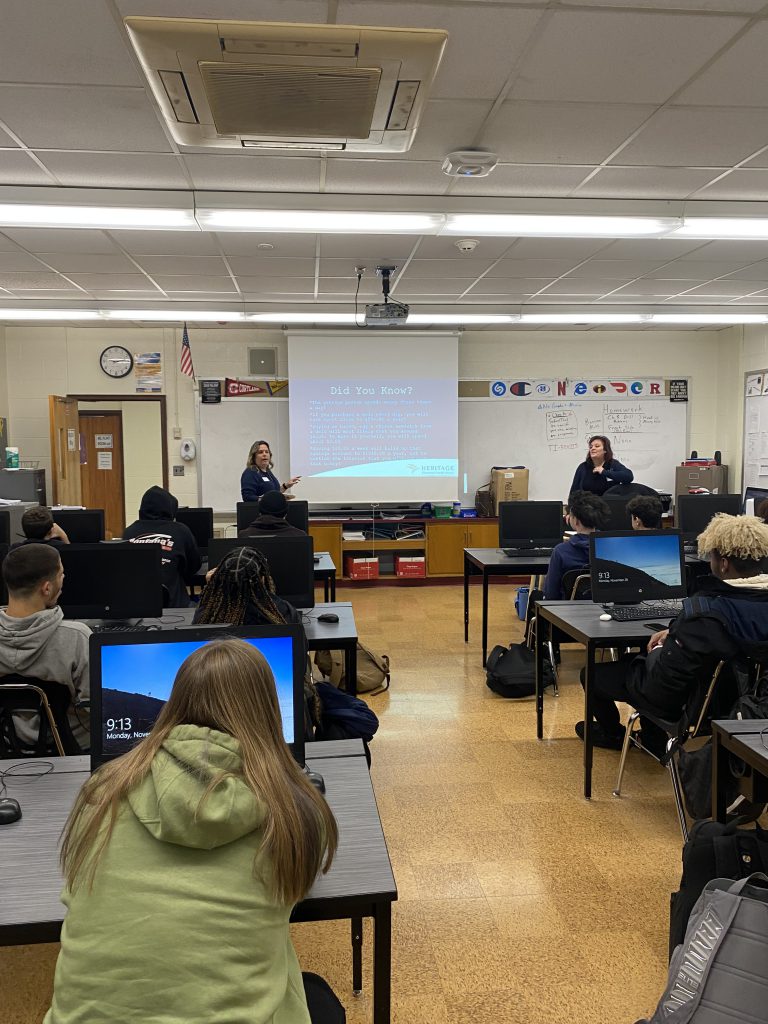 A photo taken from the back of a classroom. There are two adults presenting at the front of the class on a screen with students sitting at desks.