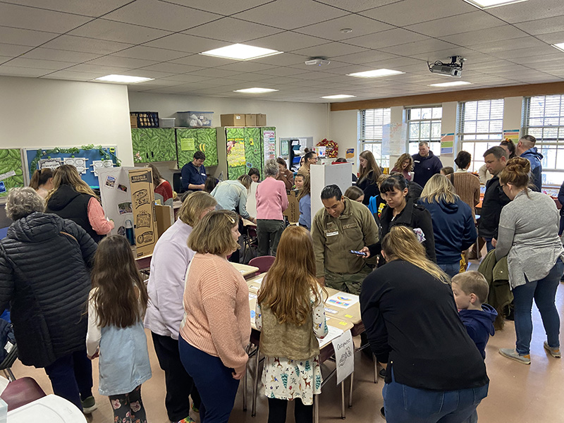 Students are at desks with their projects and many adults are walking around looking at the various projects.