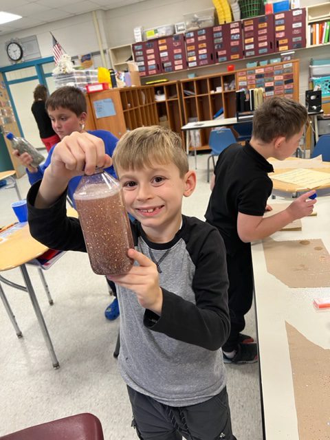 An elementary age boy, with short blonde hair and wearing a gray and black long-sleeve shirt, holds up a cup filled with liquid and glitter swirling around.