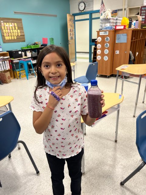 An elementary age girl, with longer dark hair and wearing a flowered shirt, holds up a cup filled with liquid and glitter swirling around.